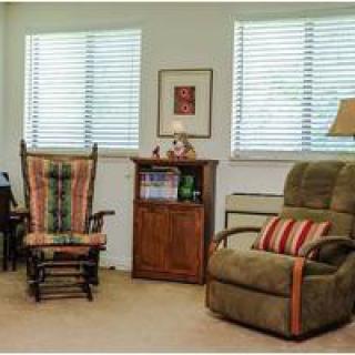 A cozy living room with a rocking chair, recliner, and a small bookshelf between two windows.