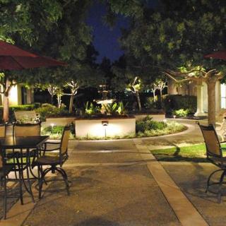 A beautifully lit patio garden in the evening, featuring tables, chairs, umbrellas, and a central fountain surrounded by lush landscaping.