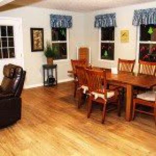 A cozy dining area with wooden floors, a large table with multiple chairs, and windows adorned with curtains.