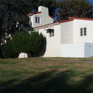 A white residential building with a red tiled roof, surrounded by green grass and trees.