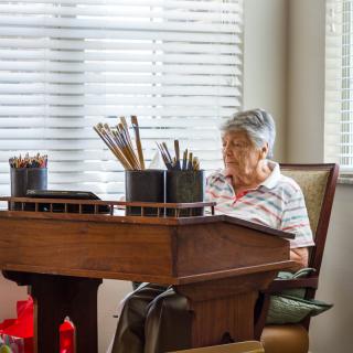 An elderly woman sits at a desk, engaging in drawing with various art supplies around her.