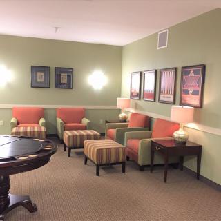 A cozy seating area with green walls, featuring armchairs, ottomans, decorative wall art, and soft lighting.
