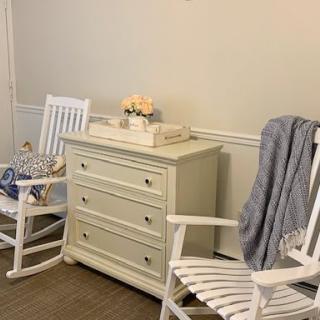 A cozy corner featuring two white rocking chairs and a dresser, decorated with a vase of flowers and a soft blanket.