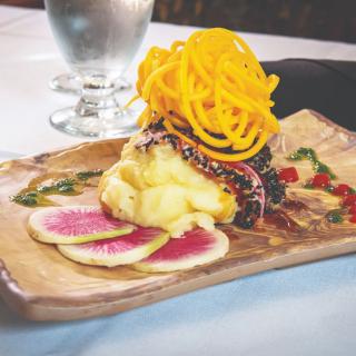 A beautifully presented gourmet dish featuring spiralized garnish atop a main component, with sliced watermelon radish and artistic sauce drizzle on a wooden platter.