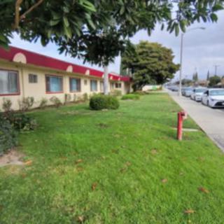 A row of buildings with red roofs next to a grassy area and sidewalk lined with parked cars on an overcast day.