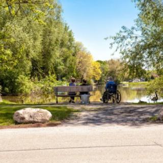 A peaceful scene in a park with people sitting on a bench by the lake, surrounded by trees.