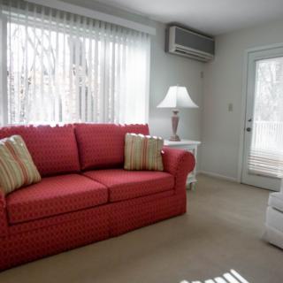 A cozy living room featuring a red sofa adorned with striped cushions, complemented by a white lamp and large windows with sheer curtains.