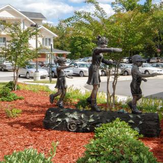 A sculpture of children playing is displayed in an outdoor garden setting with trees and shrubs.