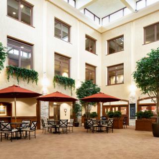 A spacious indoor atrium featuring tables and chairs under red umbrellas, surrounded by greenery and large windows.