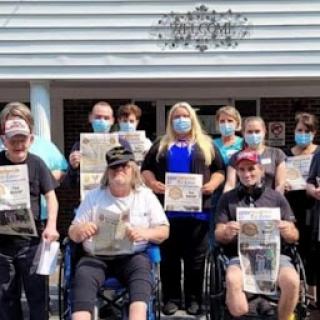 A cheerful group gathers outside holding newspapers, showcasing their camaraderie and community spirit while wearing masks.