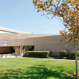 A modern building with a shaded entrance, surrounded by trees and grass on a sunny day.