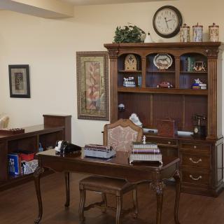 A traditional-style office with a wooden desk and bookshelf, featuring a clock and decorative items.