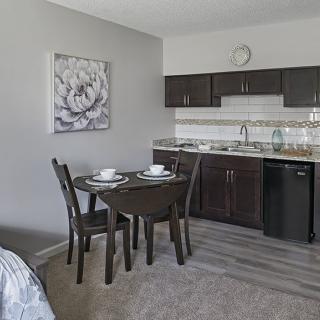 A compact kitchenette with dark cabinets and a small dining area featuring a round table and chairs, complemented by floral artwork on the wall.