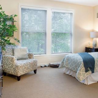 A cozy bedroom with a patterned chair, a comfortable bed, large windows with blinds, and a potted plant.