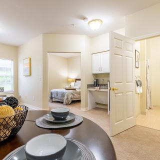 A bright and cozy apartment interior with a dining table set, a view into the bedroom, and a small kitchenette.