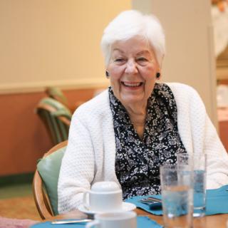 An elderly woman smiling while sitting at a dining table indoors.