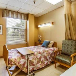 A cozy and well-lit patient room in a hospital, featuring a bed, an armchair, and natural light from the window.