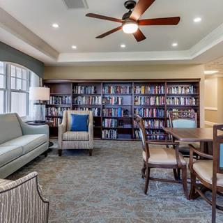 A cozy library room featuring bookshelves, comfortable seating with a sofa and armchair, and a round table under a ceiling fan.