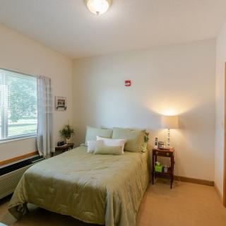 A cozy bedroom with a neatly made bed, side table, and a lamp by the window.