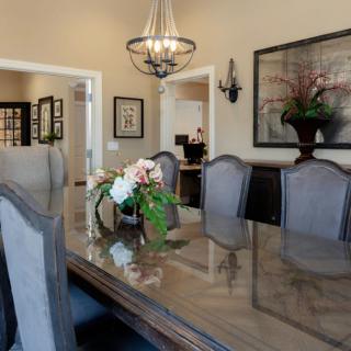 A formal dining room with a glass-top table and elegant chairs, accented by a chandelier and floral centerpiece.