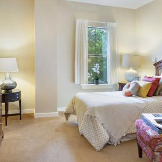 A cozy bedroom features a bed with decorative pillows, lamps on nightstands, and a window letting in natural light.
