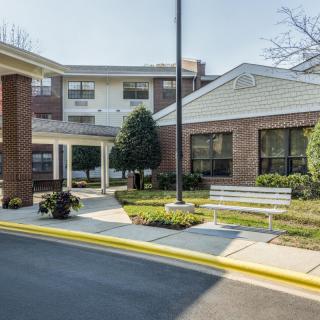 The image shows the entrance to a building surrounded by landscaping, with benches and a pathway leading to the front door.