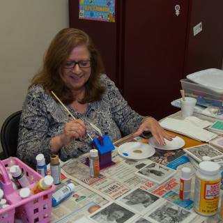 A woman is engaged in painting a small wooden house, surrounded by various art supplies.