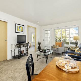 A cozy living room featuring a television, sofa, and a decorated table with natural light coming in through the windows.