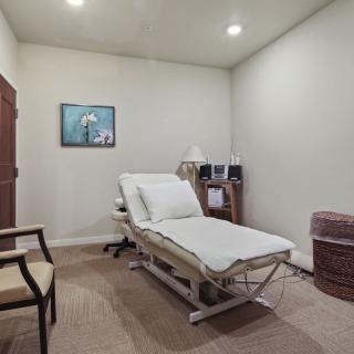 A simple and clean medical examination room with a treatment bed, chair, lamp, and wall art.