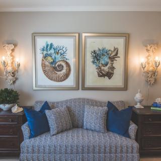 A cozy living room setup featuring a patterned sofa, blue cushions, and sea-themed artwork on the wall, illuminated by elegant sconces.