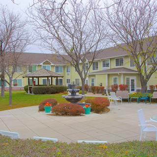 An outdoor patio area with a central fountain, surrounded by trees and benches, adjacent to a two-story building.