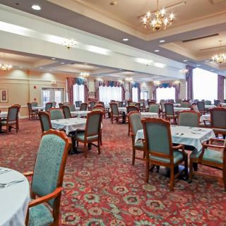 A spacious dining room with elegant chandeliers, neatly arranged tables and chairs, and a carpeted floor.