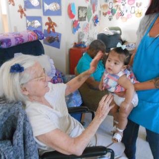 A heartwarming moment between an elderly woman and a baby, with a caregiver nearby, in a vibrant and colorful setting.
