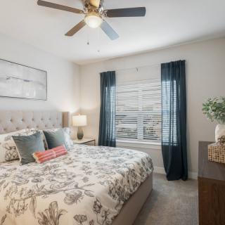 A modern bedroom with stylish decor, featuring a ceiling fan and natural light from a large window with dark curtains.