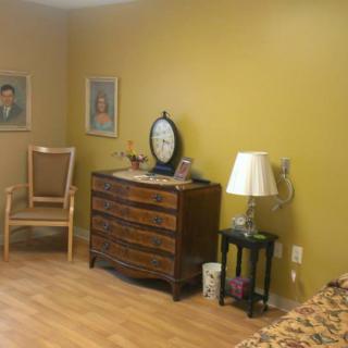 A cozy bedroom with a wooden dresser, chair, and artwork on mustard-colored walls.
