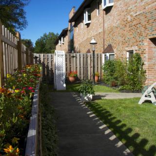 A garden with a pathway, bordered by flowers and a wooden fence, next to a brick building.