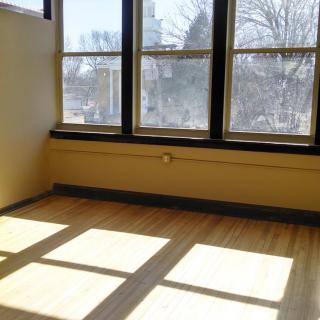 An empty room with large windows allowing sunlight to stream onto the hardwood floor.