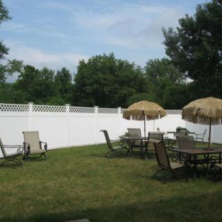 A backyard with patio furniture and sun umbrellas on a grassy lawn, surrounded by a white fence.
