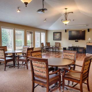 A cozy indoor dining area with multiple tables and chairs, large windows, a wall-mounted television, and stylish lighting fixtures.