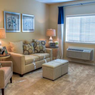 A cozy living room with a sofa, armchair, two lamps, and an ottoman. The space is well-lit by a window with blinds and curtains.