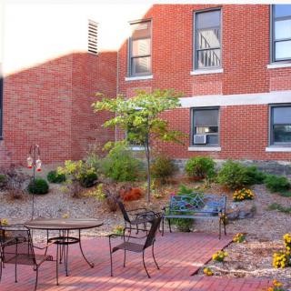A quaint outdoor courtyard with tables and chairs, surrounded by a landscaped garden adjacent to a brick building.