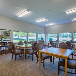 A quiet library room with tables and chairs, surrounded by bookshelves and large windows.