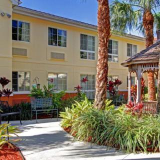 A sunny outdoor garden area with palm trees and a gazebo in front of a two-story building.