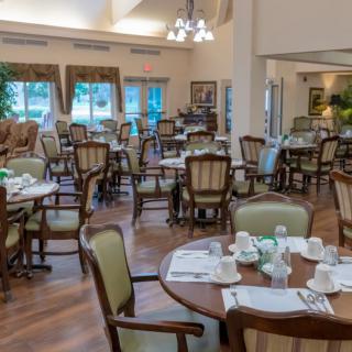 A spacious dining room with elegantly set tables and chairs, featuring large windows and chandeliers.