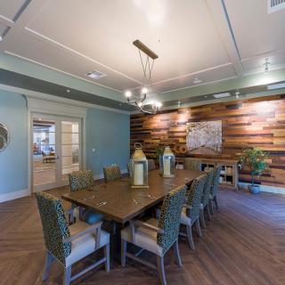 A modern dining room featuring a long wooden table, surrounded by chairs with cushions, and an accent wall of wood paneling highlighted by a chandelier.