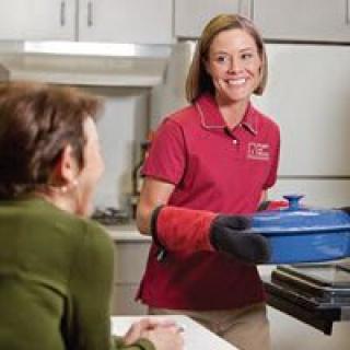 A woman wearing a red polo shirt and oven mitts is holding a blue casserole dish, smiling at a man seated nearby in the kitchen.