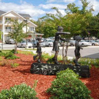 A sculpture of three children stands amidst landscaped greenery near a parking lot.