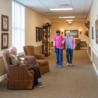 A senior care facility hallway with an elderly person sitting in a chair and two individuals walking together.
