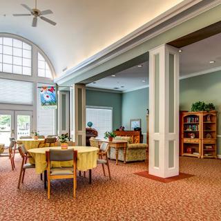 A spacious community living area featuring a dining setup with round tables and chairs, a cozy seating area with sofas, and large windows providing ample natural light.