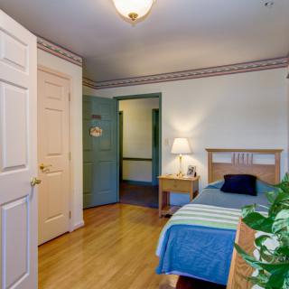 A cozy bedroom with a single bed, nightstand, and lamp, featuring wooden flooring and decorative elements.